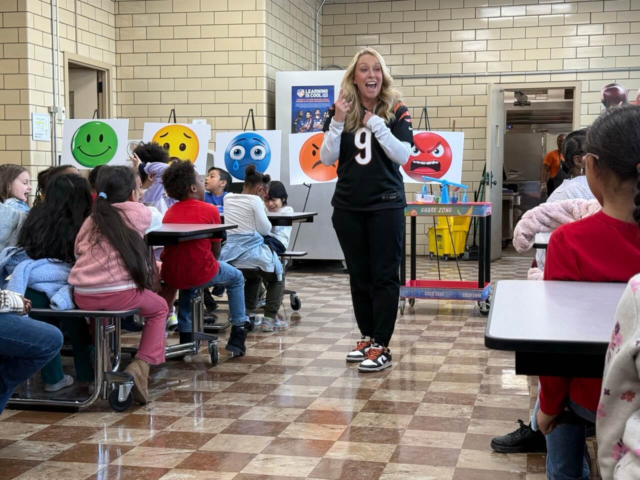 Robin Burrow smiling while giving an engaging presentation about regulating emotions to young students in the school cafeteria. Behind her are posters with various smiley faces showing all the range of emotions.